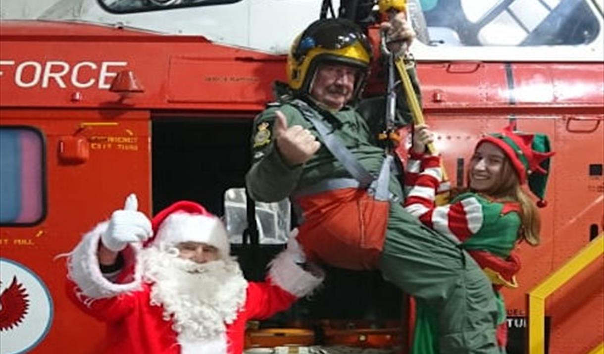 Father Christmas, an elf and helicopter pilot in a harness next to a red helicopter at the Weston-super-Mare Helicopter Museum