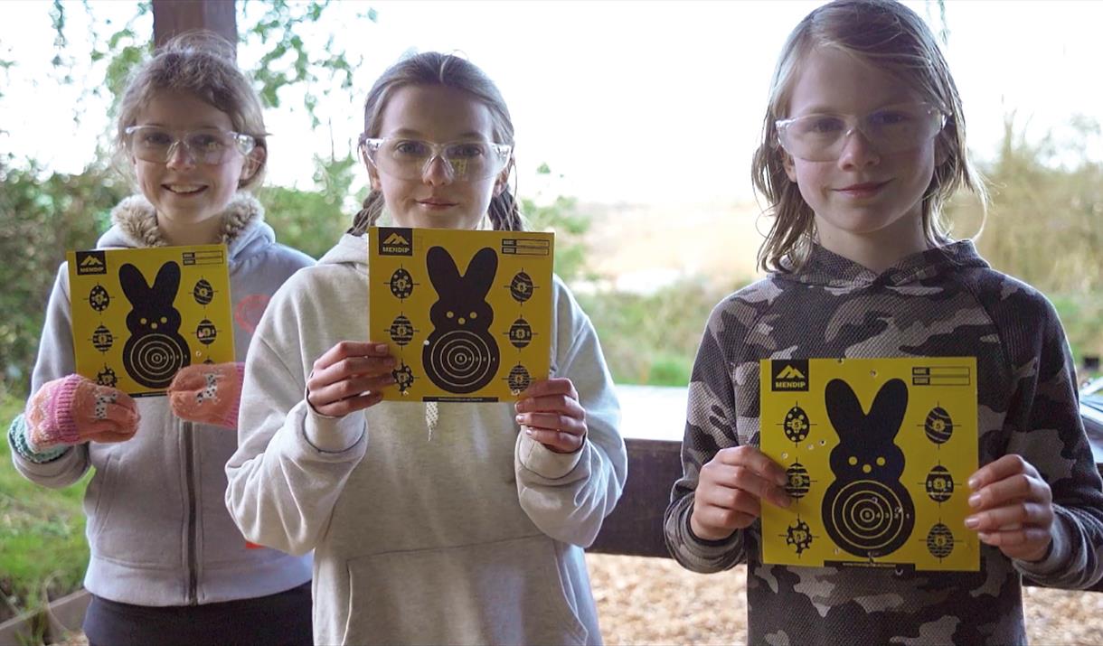 A group of children wearing protective glasses, holding up bright yellow Easter themed air rifle targets