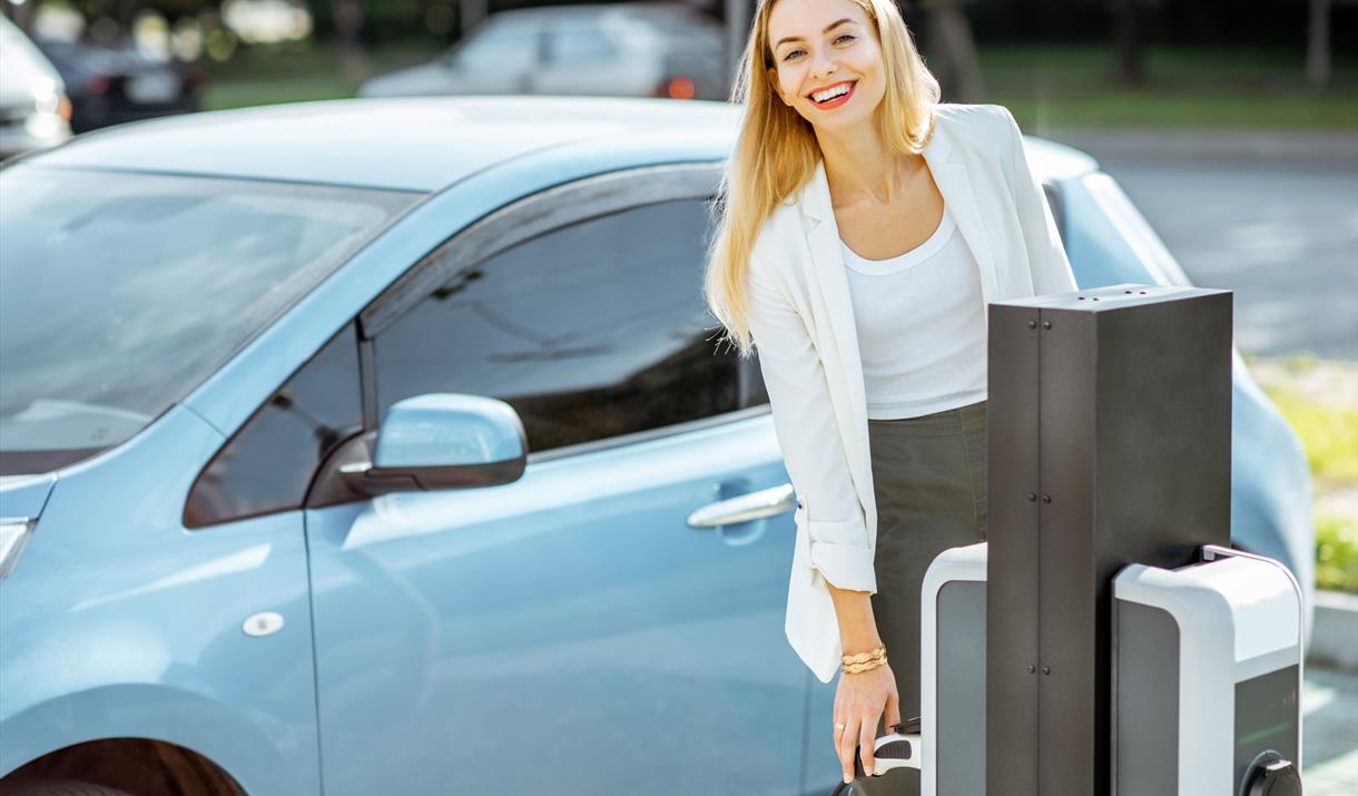 A blonde lady reaching for an EV charger to charge her blue car