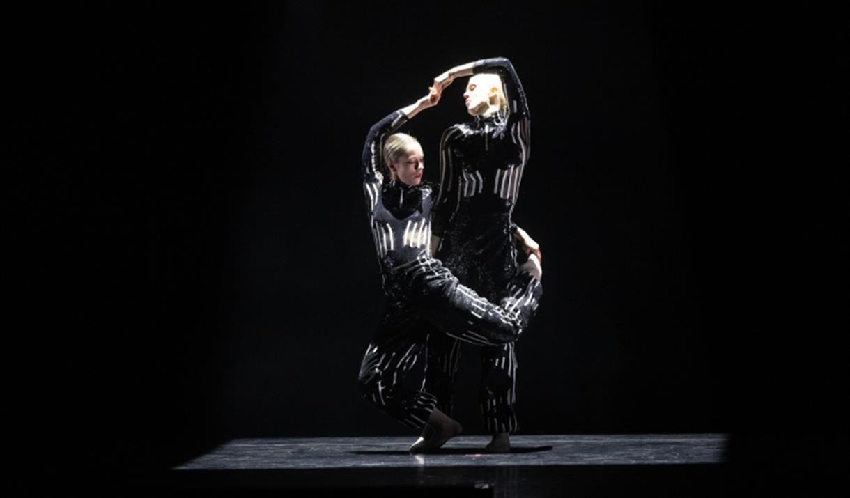 2 dancers, one with leg hooked around the other. Black background, black and white costumes