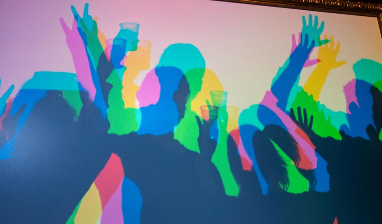 Colourful silhouettes of people dancing and holding drinks, cast against a vibrant, multi-coloured light background.