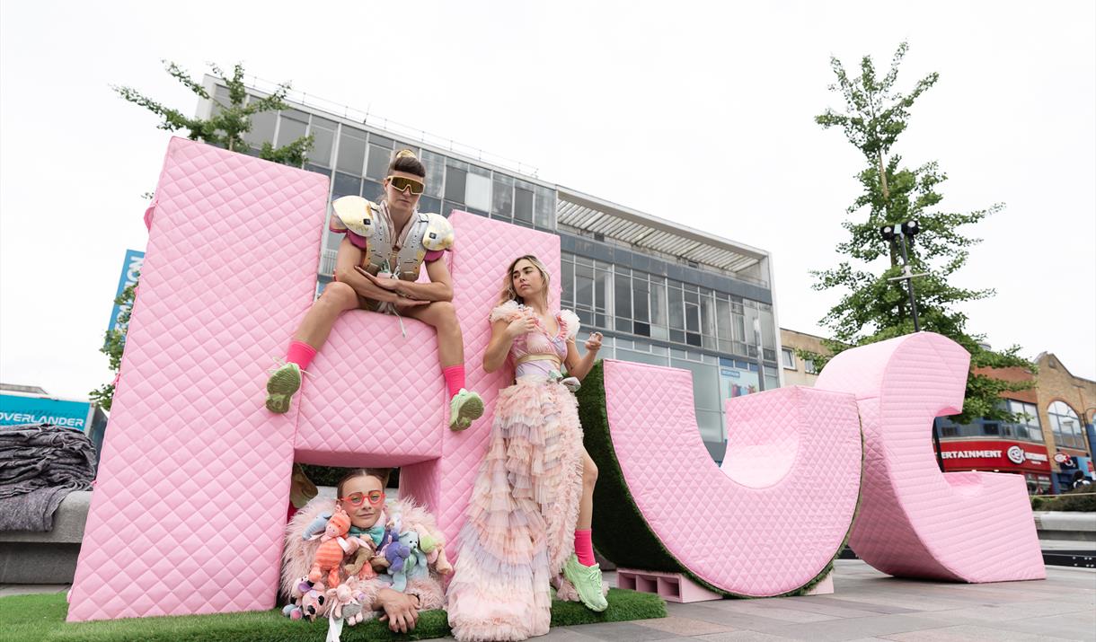 The word 'HUG' in very large pink lettering in a street. 3 women drape themselves around the H
