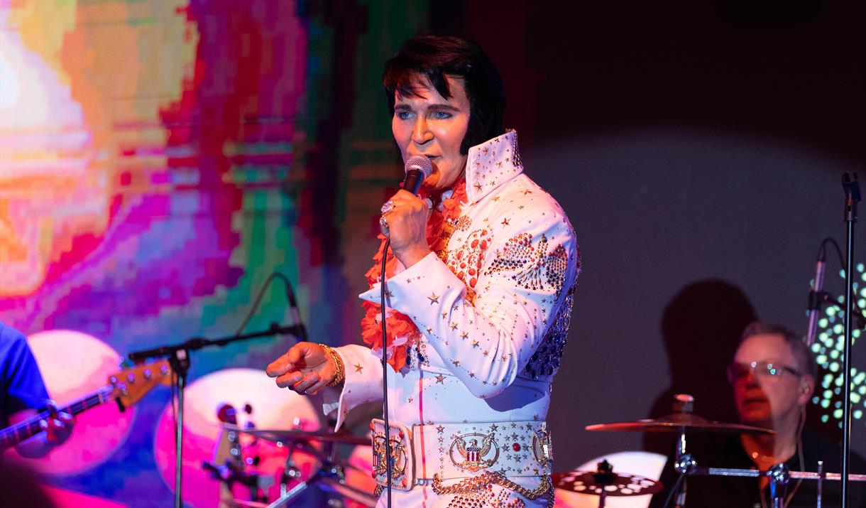 An Elvis impersonator dressed in the iconic white Elvis suit on stage and performing at the annual Harbor Lights Elvis Festival in Weston-super-Mare