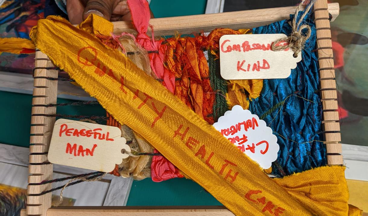 Different coloured fabrics are being woven together over a wooden frame. There are words like compassionate and peaceful attached on labels