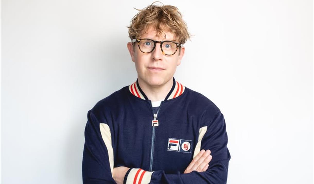 Comedian Josh Widdicombe wearing spectacles and a blue, white and red tracksuit top facing and looking straight at the camera as he is positioned in f