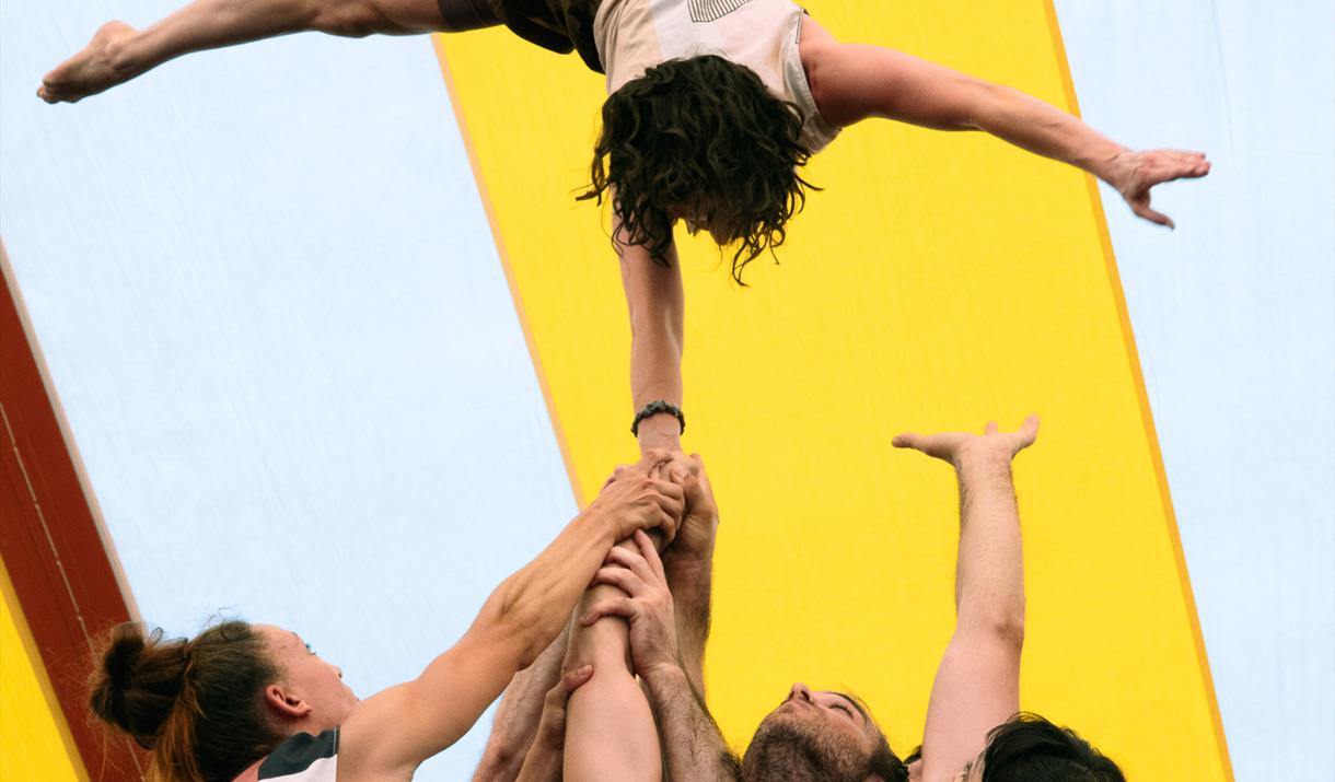 4 circus performers reach their hands up to support another performer who is resting their hands on theirs, upside down