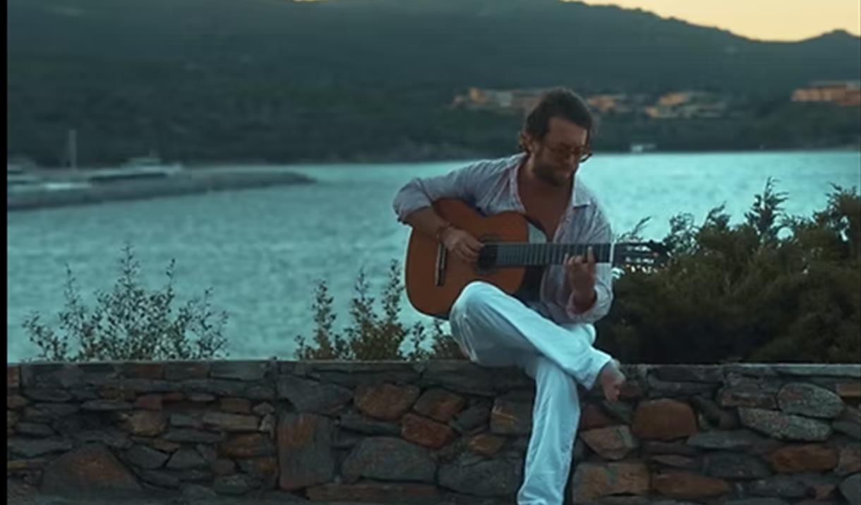 A man sitting on a wall in front of a lake playing a guitar