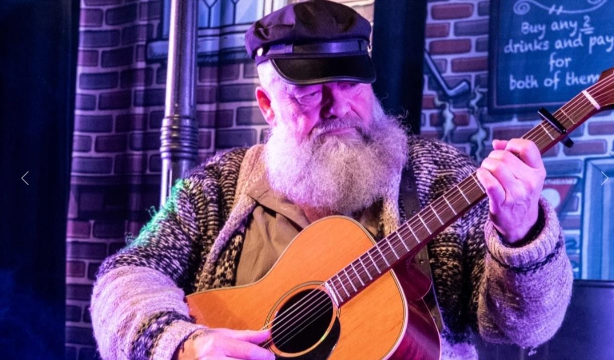 White-bearded guitarist playing his instrument against a brick wall