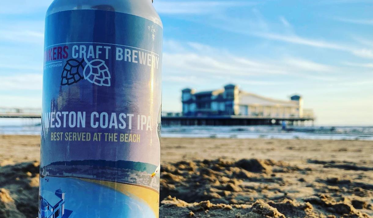 A  can of beer placed on a sandy beach with the sea and a pier in the background