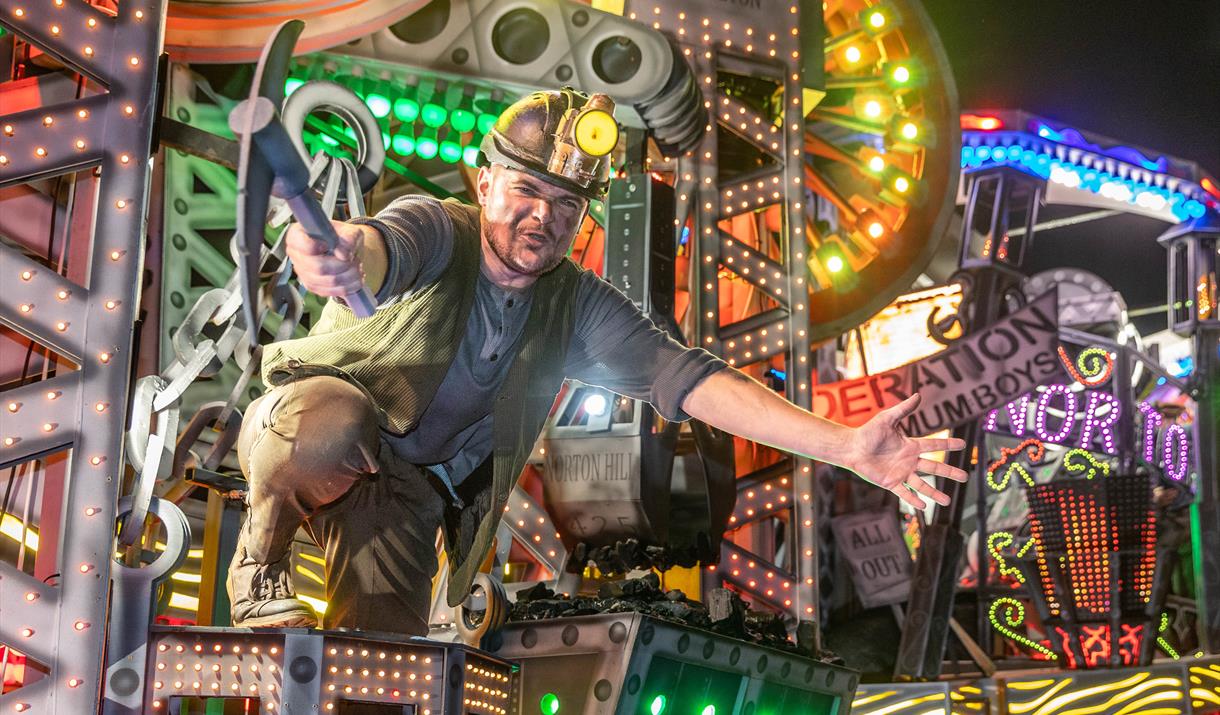 A performer in a miner's helmet and with a mining pick axe on board a brightly coloured illuminated float at the Weston-super-Mare Carnival