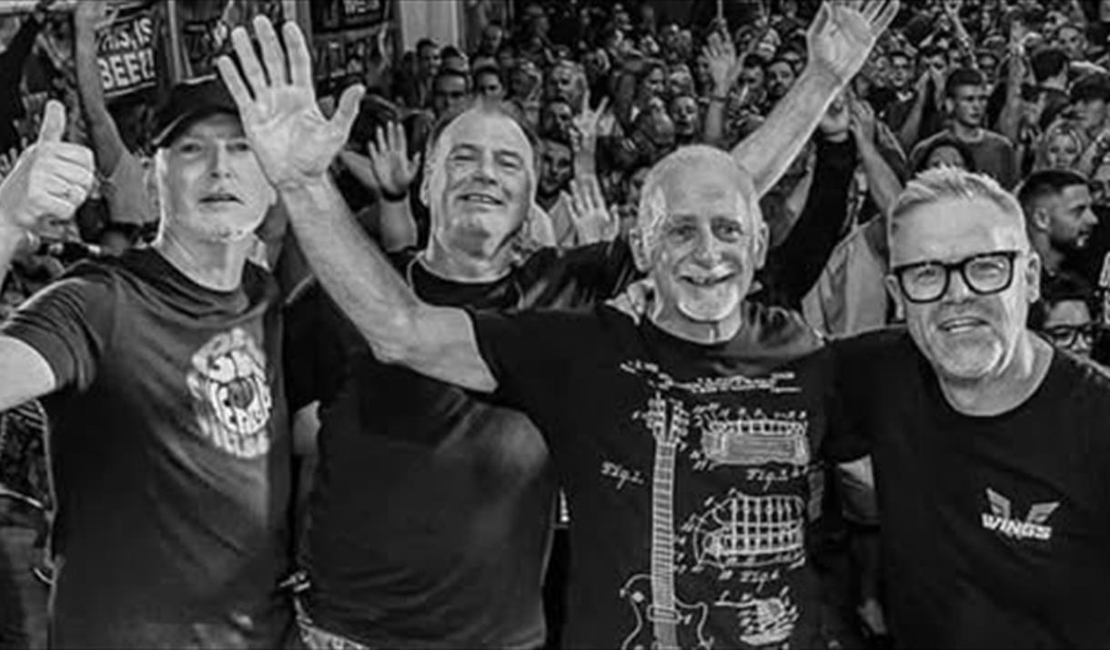 Black and white photo of four band members waving at the camera with a large crowd behind