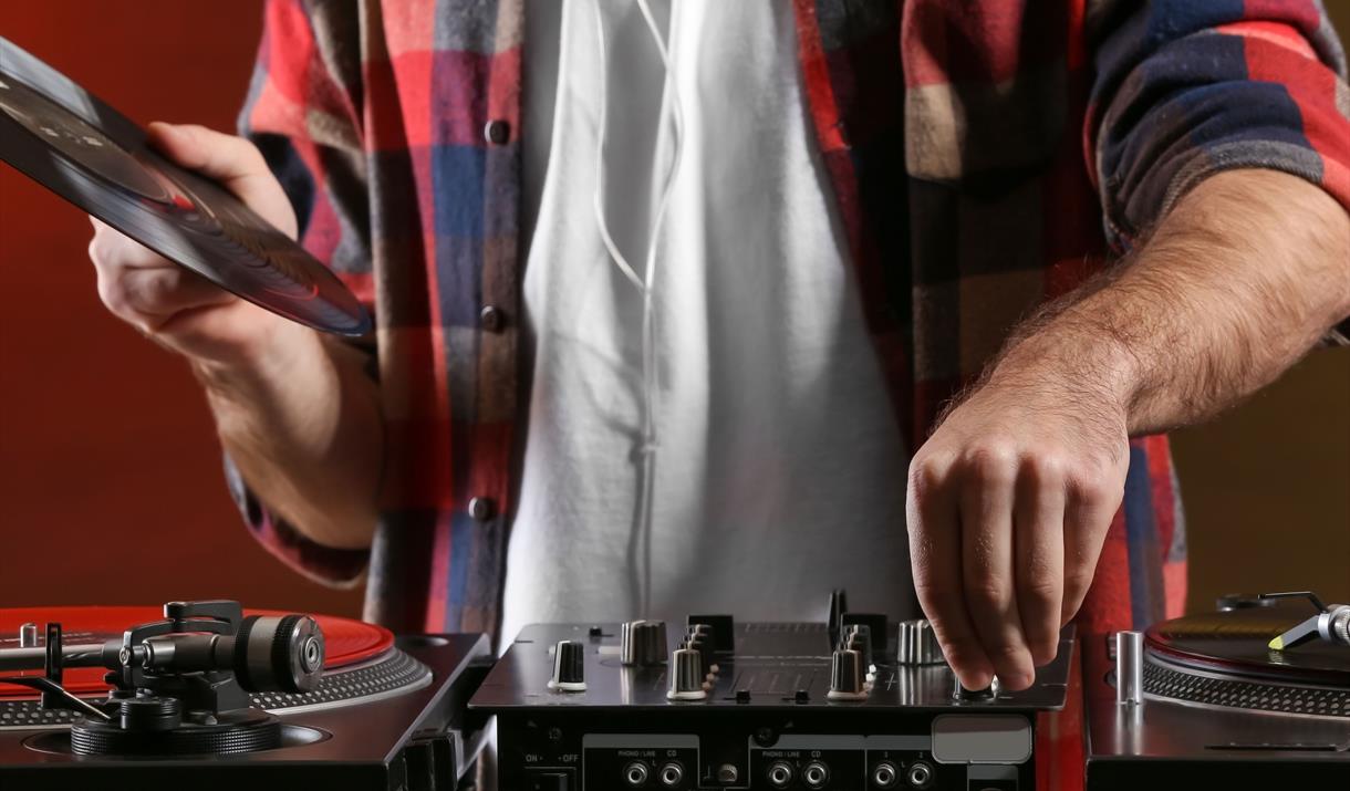 A man in a checked shirt and white undershirt (cropped without a head) holding a record above some decks