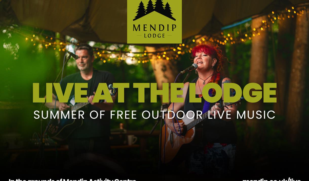 Man and woman (with red hair) performing (singing/playing guitar) under a parachute canopy, decorated with fairy lights. Top of image shows Mendip Lod