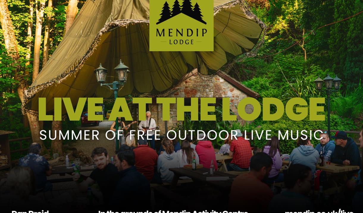 Two musicians (man and woman) playing guitar and singing into microphone under tented woodland canopy. People sat on picnic benches in front of Mendip