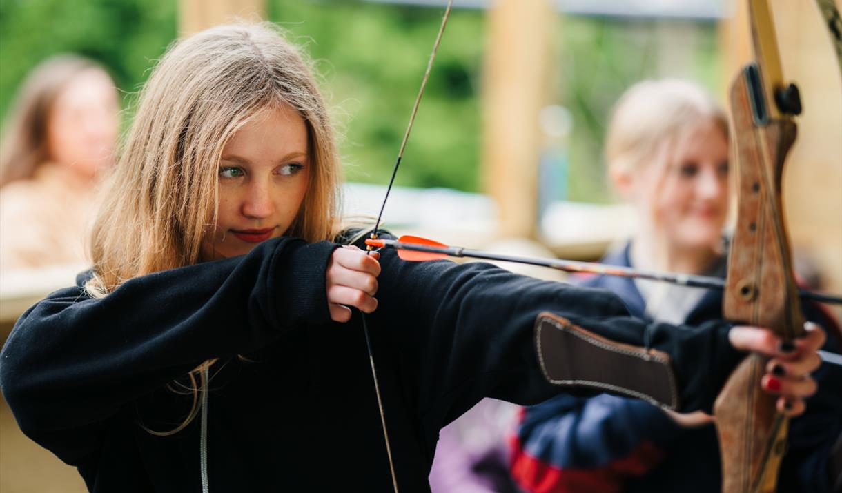 Teenage girl with blonde hair, wearing a black hoodie, pulling back an arrow on an archery bow. Looking straight down the arrow with determination.