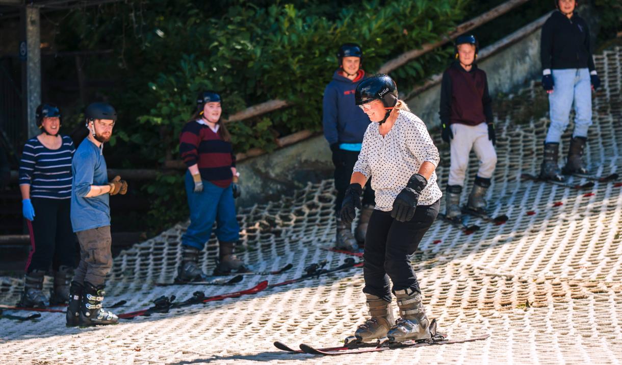 Middle-aged lady wearing skis, gloves and a helmet, looking forward as she glides along the bottom of the ski slope. In the background a Mendip Activi