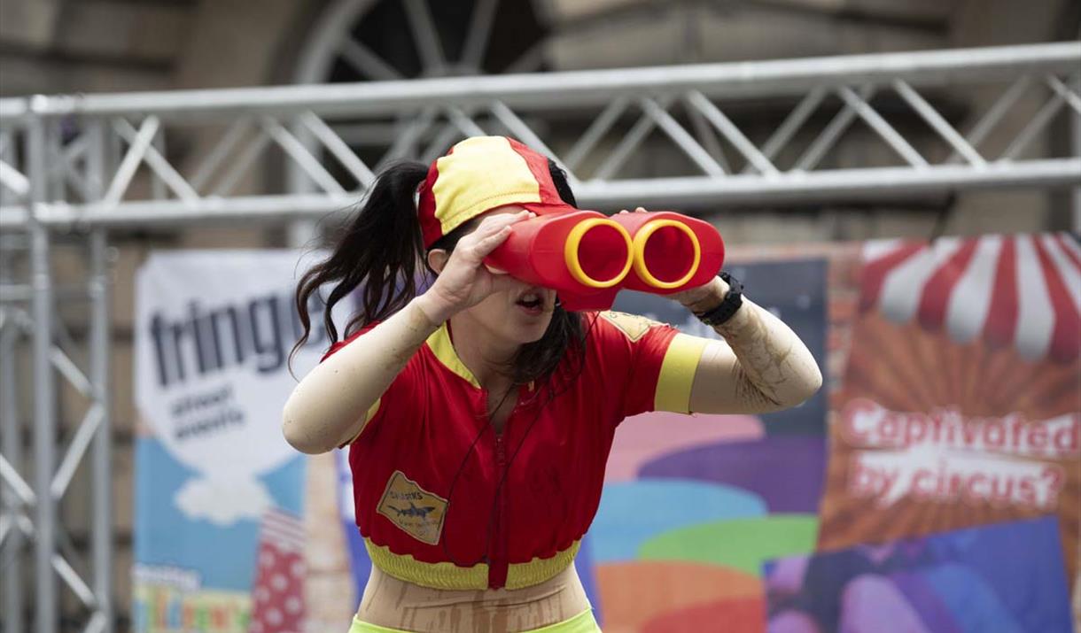 A lady in a red and yellow lifeguard costume staring through an over-large pair of binoculars