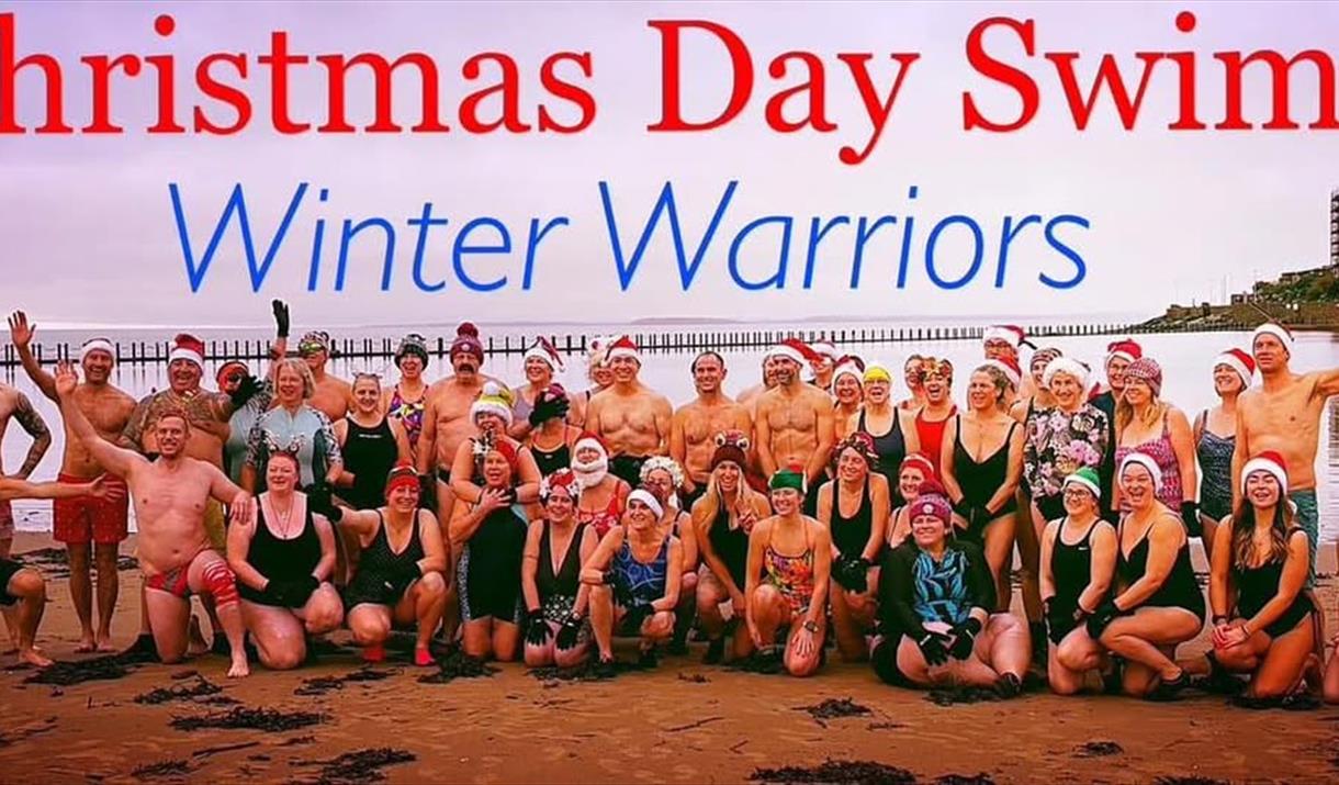 A large group of swimmers standing in front of Marine Lake Weston-super-Mare under a headline promoting the Winter Warriors Christmas Day Swim