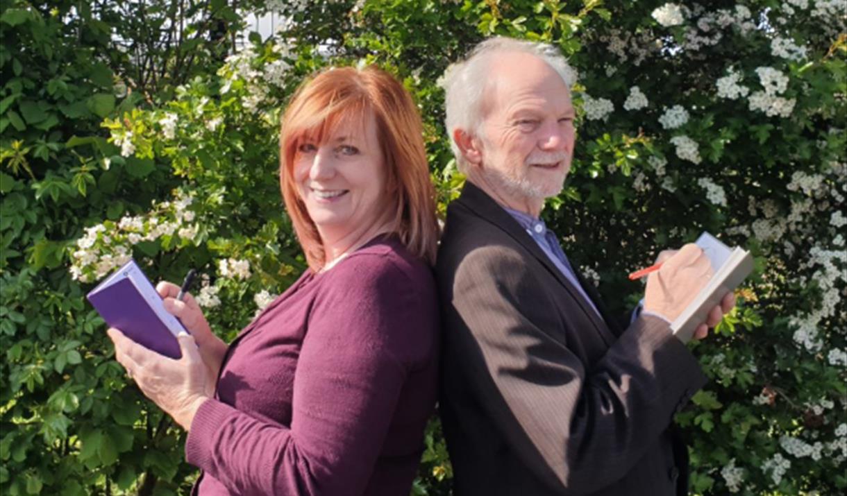 Two people standing back-to-back holding books. The background is a bush with white flowers.