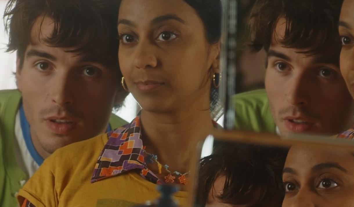 heads and shoulders of a young couple looking into mirrors so that you can see them reflected back. White male with a green jumper and and Asian femal