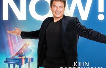 John Barrowman dressed all in black standing with his arms outstretched in front of a Grand Piano