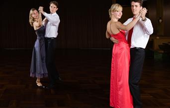 Two pairs of ballroom dancers. The men are in white shirts and black trousers and the ladies are wearing a red dress and a blue dress