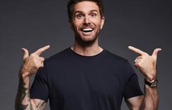 TV Presenter Joel Dommett wearing a black tee shirt is facing the camera and pointing with both hands to his smiling face