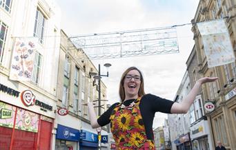 Woman with arms in air on Weston high street with two flags hanging behind her under the welcome to weston sign