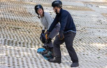 A smiling lady in a helmet on a snowboard receiving tuition from an instructor at the Mendip Activity Centre