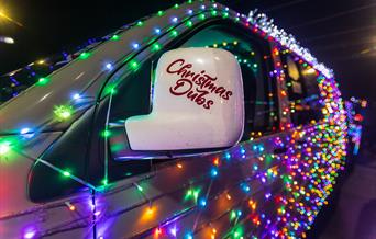 A VW van decorated in twinkling Christmas lights and the words Christmas Dubs on the wing mirror promoting the Vee Dub Christmas Cruz drives around We