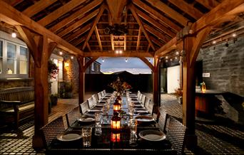A large dining area under wooden beams which opens out to the evening sky. The large table at this Webbington Farm Holiday cottage is lit with lanters