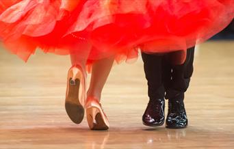 photo showing the lower legs and feet of ballroom dancers on a wooden dancefloor. She is wearing a red dress and gold shoes and he is wearing black tr