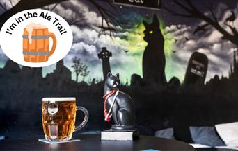 A pint of beer and a black cat set against a mural featuring a black cat and over-stamped with an I'm in the ale trail logo