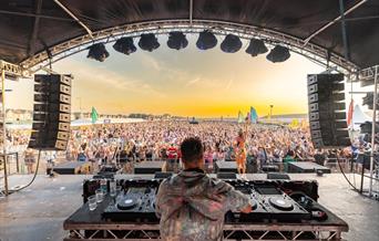 View from behind a DJ on his decks looking out across the stage onto a huge crowd at an outdoor music festival