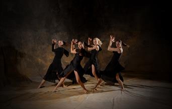 Four female dancers in flowing black dresses perform a synchronised routine on a dimly lit stage with a textured, earthy backdrop. They strike dramati