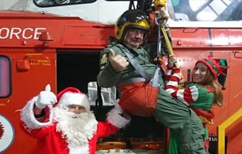 Father Christmas, an elf and helicopter pilot in a harness next to a red helicopter at the Weston-super-Mare Helicopter Museum