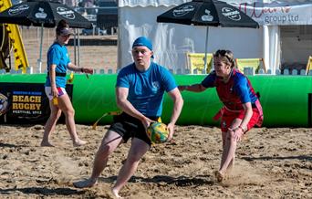 Beach rugby player in blue kit about to throw the ball before being tackled by another player