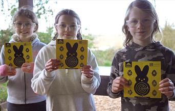 A group of children wearing protective glasses, holding up bright yellow Easter themed air rifle targets