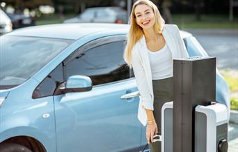A blonde lady reaching for an EV charger to charge her blue car