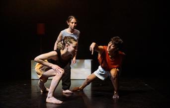 2 people crouching and communicating in dance while another stands behind observing. On a stage, dimmed lighting