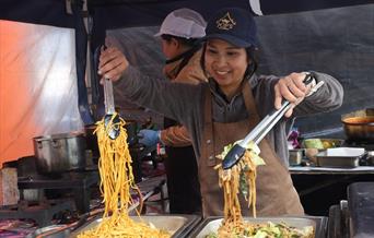 A lady stall holder at the Eat Weston Food Festival holding a tong full of noodles and another full of vegetables over two hot food platters