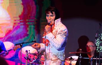An Elvis impersonator dressed in the iconic white Elvis suit on stage and performing at the annual Harbor Lights Elvis Festival in Weston-super-Mare