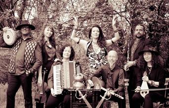 Black and white image of seven people with instruments standing in a wooded area and smiling to camera