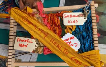 Different coloured fabrics are being woven together over a wooden frame. There are words like compassionate and peaceful attached on labels