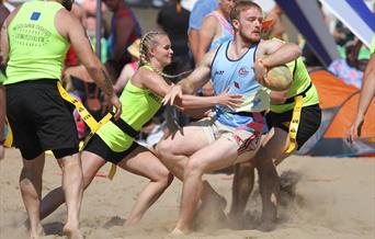 A woman in yellow attempts to tackle a man in blue during a Beach Rugby Festival