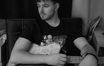 Black and white image of Luke Potter sat with an electric guitar and event information.