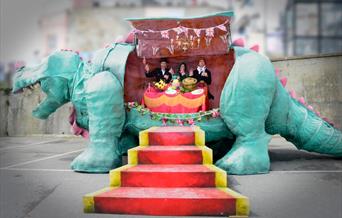 A model dinosaur cut open and inside its belly are three people sitting at a dining table eating
