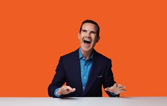 Photograph of Jimmy Carr stood against a orange background.