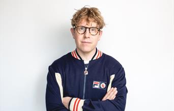 Comedian Josh Widdicombe wearing spectacles and a blue, white and red tracksuit top facing and looking straight at the camera as he is positioned in f