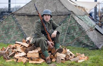 A soldier in WW2 uniform holding a rifle and sitting on a pile of split logs at the Weston-super-Mare Helicopter Museum Military History weekend